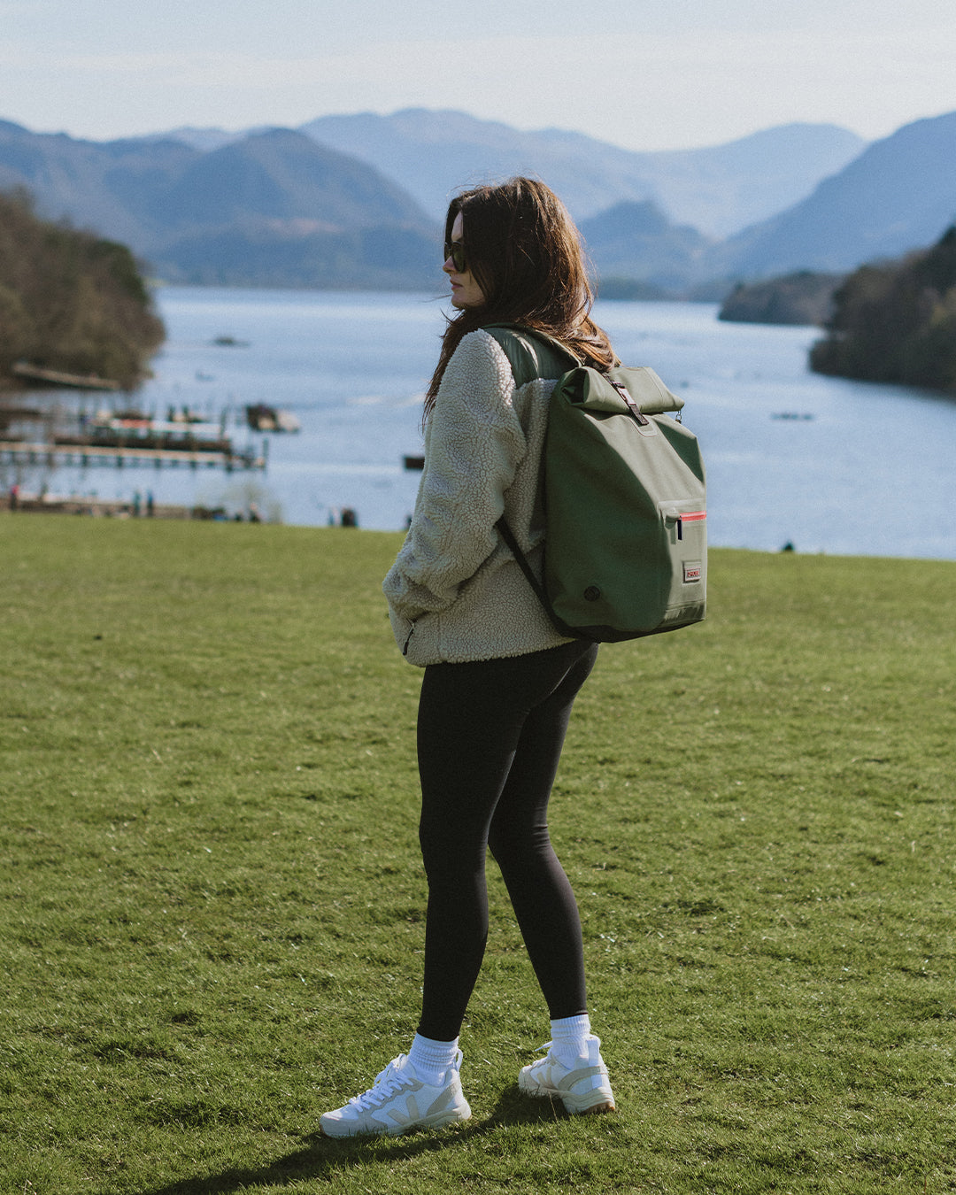 Female model walking in the hills of the lake district wearing our stylish, multi-functional moss green roll-top rucksacks, which have extra storage and waterproof outer for all-weather protection. Perfect as an after swim coat, surf robe, beach coat and dog walking jacket. 